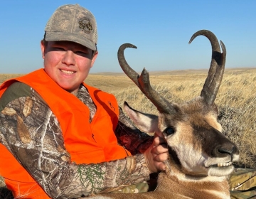 A young hunter, equipped with a blaze orange vest, smiles while presenting their first antelope trophy, commemorating a successful outing guided by SNS Outfitter & Guides.